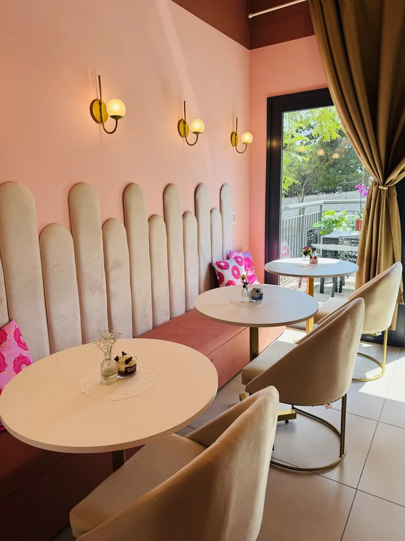 Salão com banco estofado em gomos creme, parede rosa, arandelas douradas e mesas redondas brancas